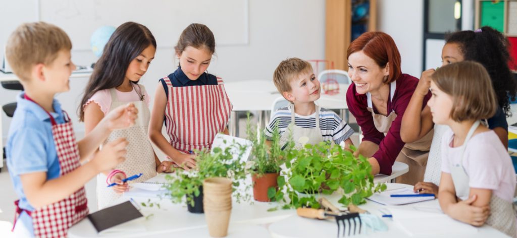 Eine Gruppe Grundschulkinder steht mit ihrer Lehrerin in einem Kreis um einen Tisch herum und pflanzt Kräuter
