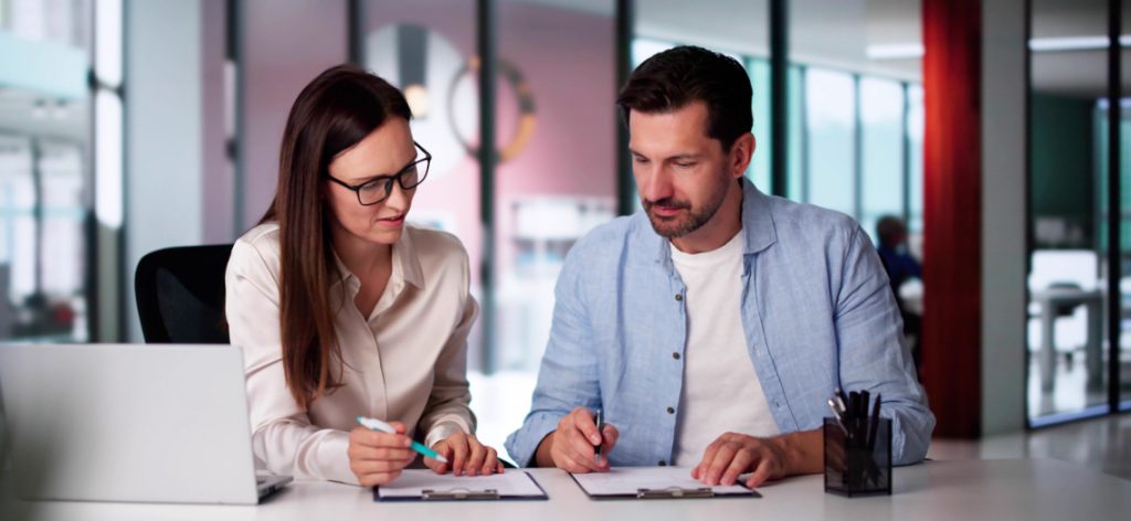 Eine Frau und ein Mann in Business Casual Kleidung in einem Büro mit Veträgen und Laptop an einem Tisch