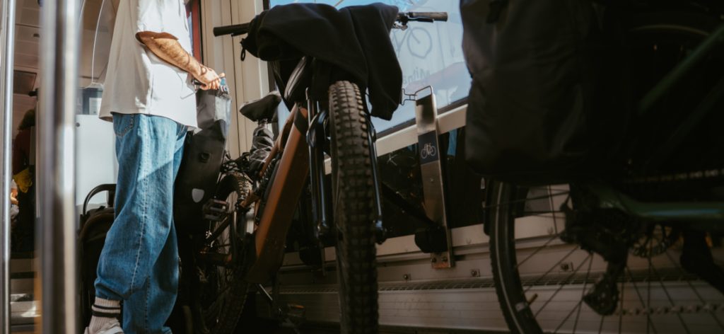 A young man standing inside a train with his bicycle, taking something from a bag attached to the bike while looking out the train window. The image shows a moment of multimodal commuting, combining cycling and public transportation during a train journey