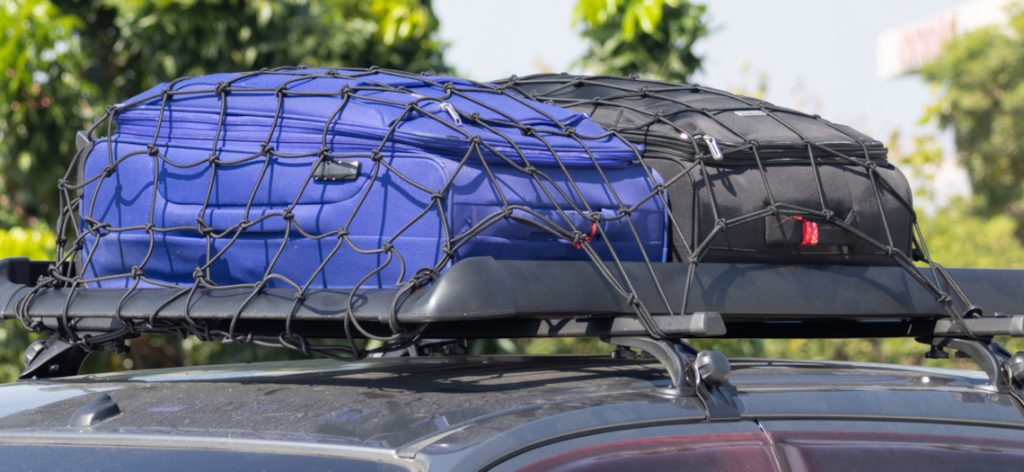 Car roof rack holds blue and black cargo nets. Gear secured for travel or outdoor adventure. Bright daylight, green trees in background. Ideal for road trip, camping, or luggage transport visuals