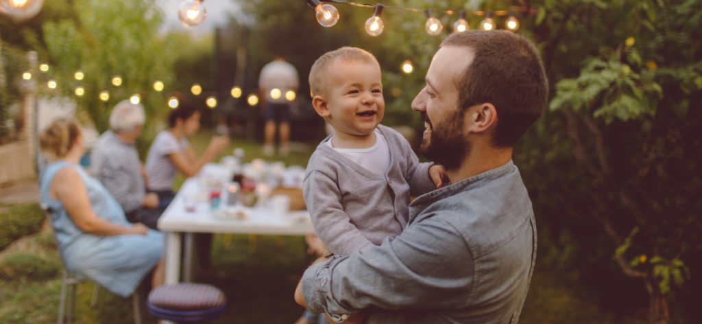 Photo of a multi-generation family having dinner outdoors in their back yard, while their little boy is celebrating his birthday